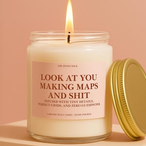 May include: A clear glass jar candle with a lit wick and a gold metal lid. The label is light pink with dark brown text that reads: THE PEONY WICK, LOOK AT YOU MAKING MAPS AND SHIT, INFUSED WITH TINY DETAILS, PERFECT GRIDS, AND ZERO GUESSWORK, 100% SOY WAX CANDLE | HAND-POURED.