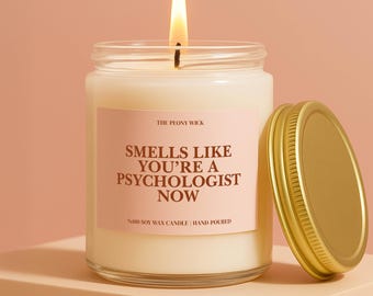 May include: A burning candle in a glass jar with a gold lid. The label says "SMELLS LIKE YOU'RE A PSYCHOLOGIST NOW." The candle is made of soy wax and hand-poured. The background is a peach colour.