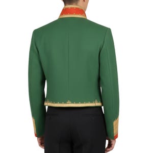 K&ouml;nnte beinhalten: Gr&uuml;ne Jacke mit goldfarbenen Verzierungen und roten Akzenten an Kragen und Manschetten. Die Jacke hat eine taillierte Passform und wird mit einer schwarzen Hose getragen. Das Design der Jacke weist verzierte goldene Stickereien an den Kanten auf.