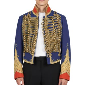 K&ouml;nnte beinhalten: Blaue Jacke im Milit&auml;rstil mit goldenen Seildetails und goldenen Kn&ouml;pfen. Die Jacke hat einen roten Kragen und Manschetten mit goldenen Verzierungen. Die &Auml;rmel haben goldene und rote Zierakzente. Die Jacke wird &uuml;ber einem wei&szlig;en Hemd getragen.