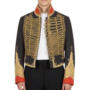K&ouml;nnte beinhalten: Schwarze Jacke im Milit&auml;rstil mit goldenen Seildetails und goldenen Kn&ouml;pfen. Die Jacke hat einen roten Kragen und Manschetten mit goldener Stickerei auf den &Auml;rmeln. Ein wei&szlig;es Hemd ist darunter sichtbar.