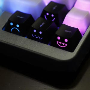 May include: Close-up of a mechanical keyboard with custom keycaps. The black keycaps feature illuminated emoji faces in blue and pink. The keyboard has a dark grey frame and white and pink keys.