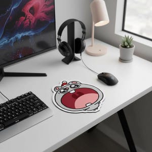 Puede incluir: Una alfombrilla de ratón de dibujos animados con un hipopótamo gris con la boca roja abierta y un gorro de Papá Noel. La alfombrilla está sobre un escritorio blanco junto a un teclado, un monitor de ordenador, unos auriculares y un ratón.