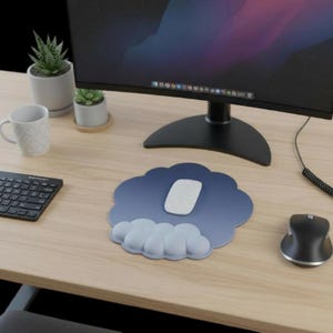 Könnte beinhalten: Ein blaues, wolkenförmiges Mauspad mit einer weißen Maus auf einem Holztisch. Das Mauspad hat eine Farbverlaufsfarbe und eine wolkenförmige Handgelenkauflage. Eine schwarze Tastatur, eine schwarze Maus, ein Computermonitor und Pflanzen befinden sich ebenfalls auf dem Schreibtisch.