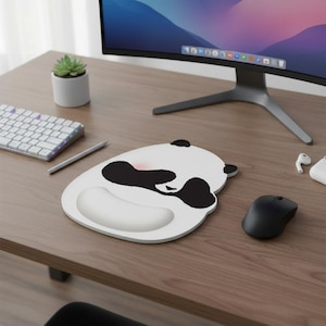 May include: A white panda-shaped mouse mat with a black and pink design, including a wrist rest. A black computer mouse is to the right of the mat. A keyboard, pen, and computer monitor are also visible.