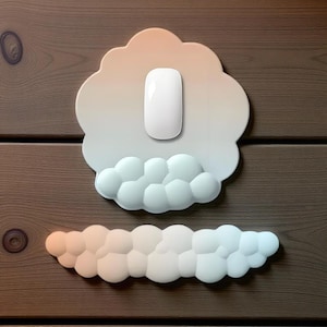 May include: A white wireless computer mouse sits on a cloud-shaped holder. Below, a matching cloud-shaped accessory is visible. The holder and accessory have a gradient colour scheme, transitioning from peach to white and blue. The items are set against a wooden background.