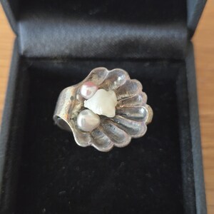 Può includere: Un anello in argento a forma di conchiglia smerlata, caratterizzato da una superficie metallica testurizzata. Il centro della conchiglia è decorato con una piccola perla rosa, una perla bianca e una pietra bianca irregolare al centro.
