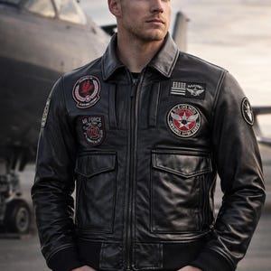 May include: Black leather jacket with multiple patches, a zipper closure, and two front pockets. The jacket has a ribbed hem and cuffs. The background features an airplane.
