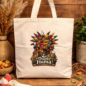 May include: Cream-colored tote bag featuring a colorful illustration of a Diablo Huma mask. The mask has a red and yellow face with a smiling expression, adorned with a headdress of various colors. The text "Diablo Huma" is below the mask.