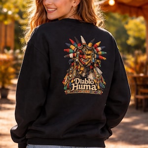 May include: Black sweatshirt featuring a colorful graphic of a tribal mask with the text "Diablo Huma." The mask design includes red, yellow, and blue accents. The sweatshirt is worn by a person in blue jean shorts.