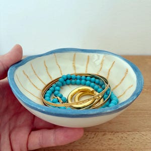 Op de afbeelding: Een kleine, ronde, gebroken witte keramische kom met een golvende blauwe rand en dunne, verticale beige strepen aan de binnenzijde. In de kom ligt een verzameling sieraden, waaronder een streng turkooiskleurige kralen en diverse goudkleurige ringen.