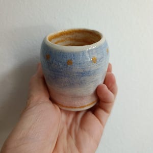 Peut inclure: Une petite tasse en céramique tournée à la main avec un dégradé de bleu clair et de blanc et une finition craquelée. L'extérieur est orné d'une rangée horizontale de petits points orange texturés, et le bord intérieur est émaillé d'orange doré.