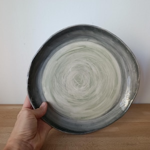 Può includere: Un piatto rotondo in ceramica fatto a mano con un ampio bordo grigio antracite scuro che sfuma verso un centro verde salvia pallido più chiaro. La superficie presenta visibili texture a pennellate vorticose, creando un motivo circolare dinamico sull'intera superficie del piatto.