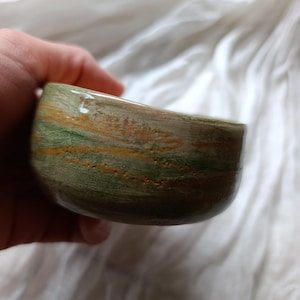 Könnte beinhalten: Eine kleine, handgefertigte Schale mit einem einzigartigen, organischen Design. Die Schale weist eine Mischung aus Grün-, Braun- und Goldtönen auf, die einen Marmoreffekt erzeugt. Die glatte Oberfläche und die abgerundete Form deuten darauf hin, dass sie aus Stein oder Keramik besteht. Die Schale wird in einer Hand gehalten.