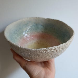 Könnte beinhalten: Eine handgefertigte Keramikschale mit einer rauen, strukturierten Außenseite und einer glatten, farbenfrohen Innenseite. Das Innere zeigt Bänder in Blau, Rosa und Gelb. Die Schale wird in einer Hand gehalten, um ihre Größe und Form zu zeigen. Ein einzigartiges Kunstwerk.