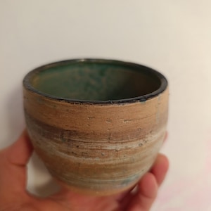 Puede incluir: Un cuenco de cerámica hecho a mano con forma redondeada. El exterior presenta bandas horizontales de color beige, marrón y blanco. El interior está esmaltado en un color verde azulado intenso. El cuenco está en una mano.