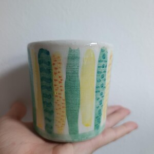 Può includere: Un bicchiere cilindrico in ceramica con base bianca caratterizzato da strisce verticali dipinte nei toni del verde e del giallo. Le strisce presentano texture variegate, tra cui pennellate piene, motivi a pois e linee ondulate.