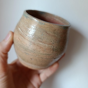 Peut inclure: Une tasse en céramique faite à la main, de forme organique unique. La tasse présente un mélange de tons terreux, notamment beige, marron et des touches de bleu sur le bord. L'intérieur est de couleur rougeâtre. La tasse est tenue dans une main sur fond blanc.