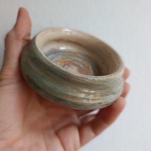 Könnte beinhalten: Eine kleine, handgefertigte Keramikschale mit runder Form. Die Außenseite weist horizontale Bänder in gedämpften Grün- und Goldtönen auf, während das Innere einen Wirbel aus Blau, Orange und Braun zeigt. Die Schale wird in einer Hand gehalten, um ihre Größe und Textur zu zeigen.