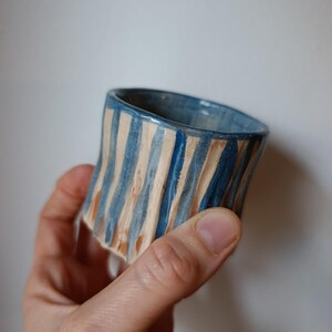 Może przedstawiać: Mały, cylindryczny kubek ceramiczny z ręcznie malowanym wzorem w pionowe niebieskie paski na jasnobeżowej, teksturowanej powierzchni gliny. Krawędź wykończona jest gładką niebieską glazurą, a na zewnątrz widoczne są pionowe żłobienia.