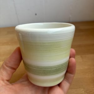 Op de afbeelding: Een kleine keramische beker met een gladde, glanzende afwerking en horizontale banden in lichtgeel, crème en saliegroen. Het object heeft een eenvoudige, ronde vorm met een licht uitlopende rand en een strak, minimalistisch uiterlijk.