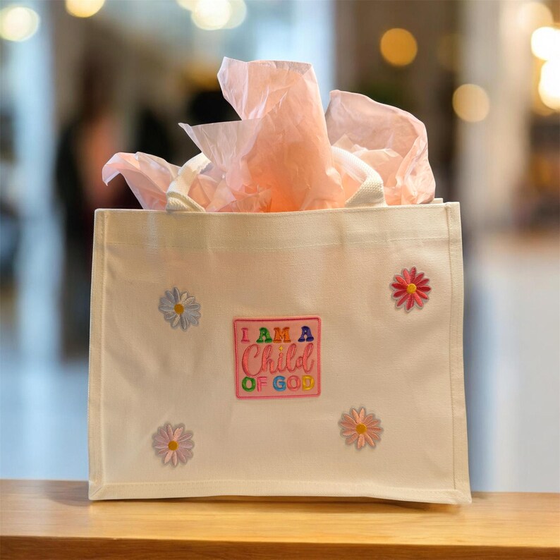 May include: A white canvas tote bag with embroidered daisy accents and a rectangular patch that reads "I AM A Child OF GOD." The bag is filled with pink tissue paper and has white handles. The bag is sitting on a wooden surface.