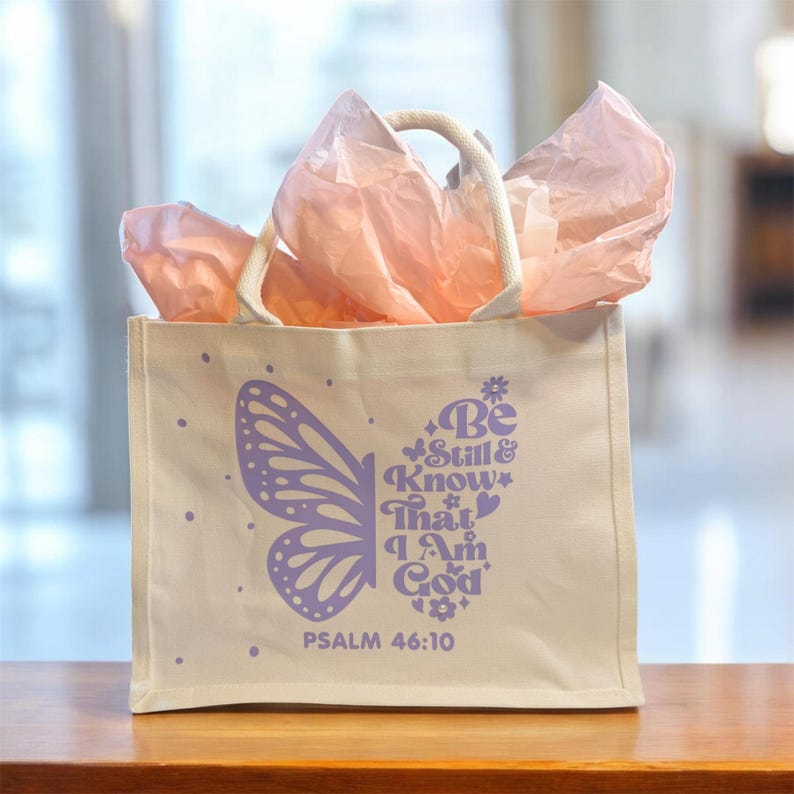 May include: A cream-colored tote bag with a butterfly wing design in lavender and the text "Be Still & Know That I Am God" and "Psalm 46:10". The bag has a white rope handle and is filled with pink tissue paper.