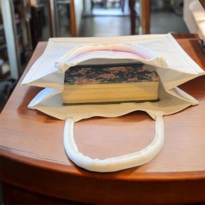 May include: A white tote bag with a rectangular shape and a rounded handle. The bag is open, revealing a thick book with a floral cover. The bag is sitting on a wooden surface.