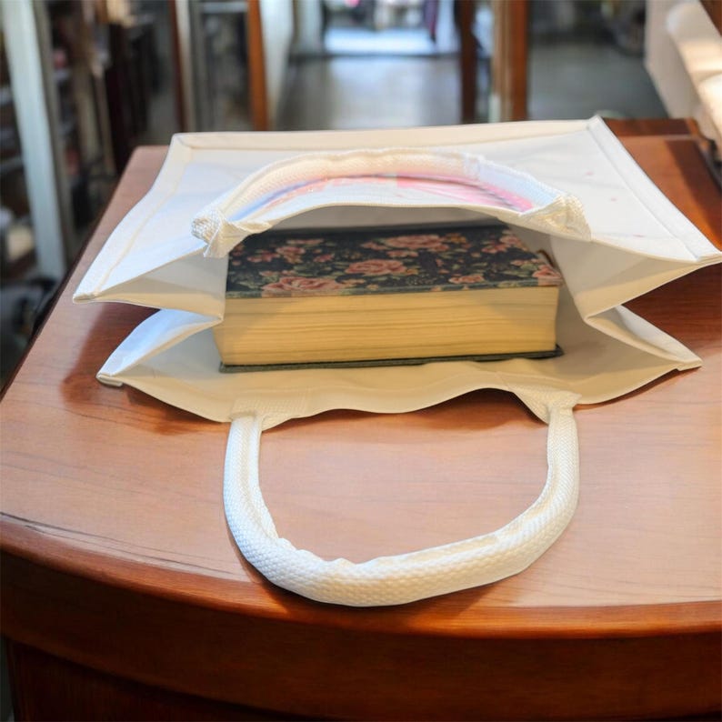 May include: A white tote bag with a rectangular shape and a rounded handle. The bag is open, revealing a thick book with a floral cover. The bag is sitting on a wooden surface.