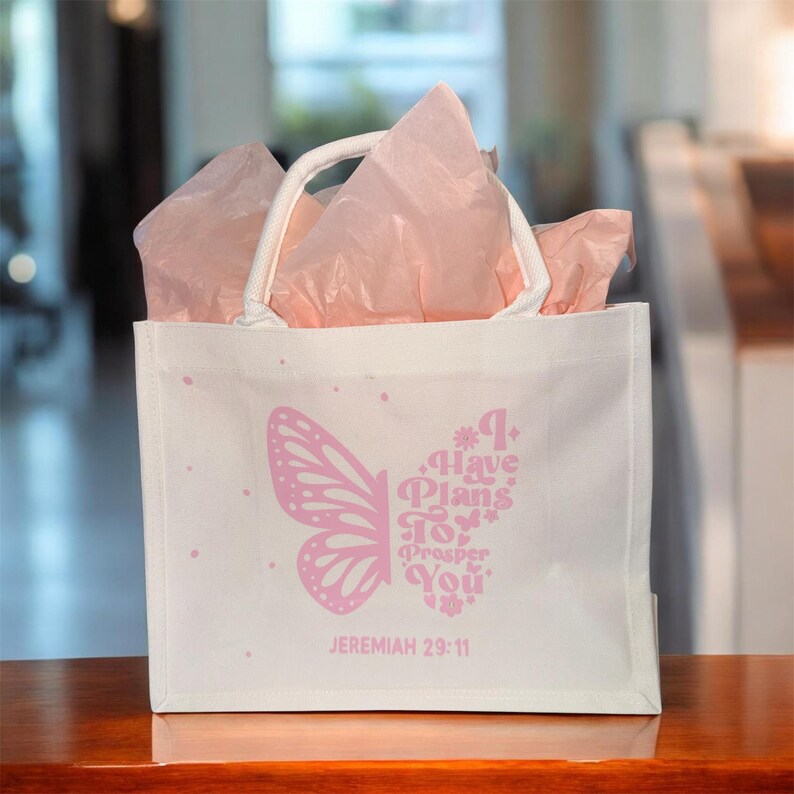 May include: White tote bag with a butterfly design and the text "I Have Plans To Prosper You" in pink. The bag contains pink tissue paper and has a white handle. The text "Jeremiah 29:11" is printed below the text.