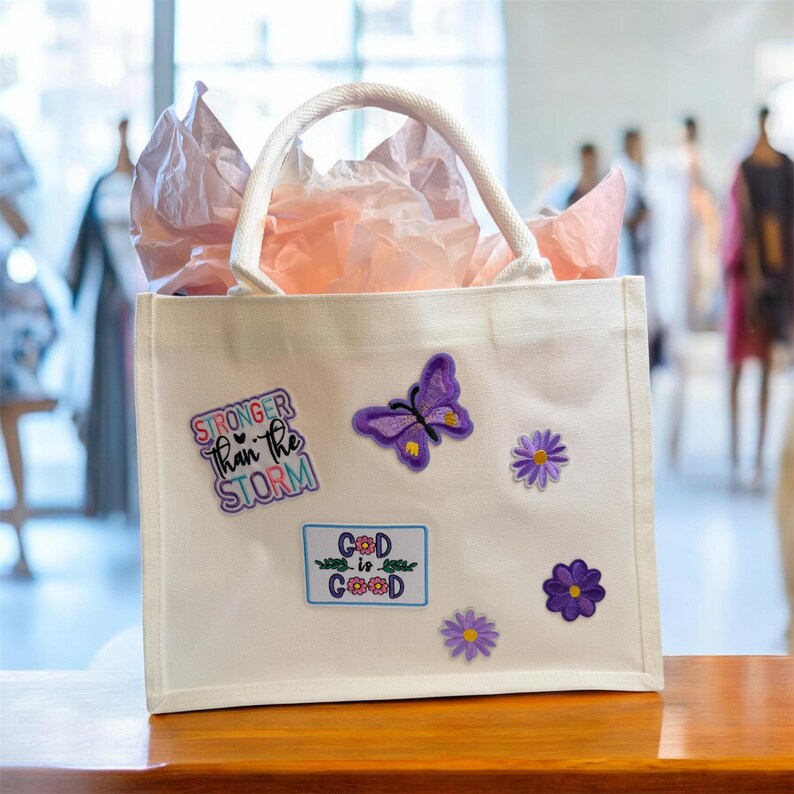 May include: A white canvas tote bag with a handle, decorated with embroidered patches. Patches include a purple butterfly, daisies, and the phrases "STRONGER Than the STORM" and "God is Good". The bag is filled with pink tissue paper.