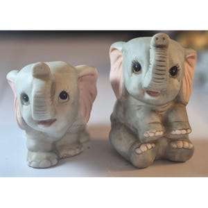 May include: Two ceramic elephant figurines. One is standing, the other is sitting. Both are light gray with pink ears and trunks. They have black eyes and smiling pink mouths. The figurines are detailed and appear to be hand-painted.