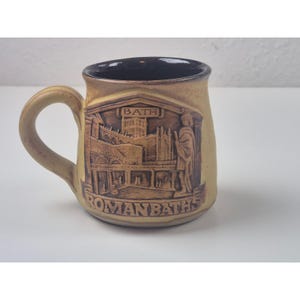 May include: A tan ceramic mug with a dark brown interior and a curved handle. The mug features a detailed relief of Roman Baths with the text "BATH" and "ROMAN BATHS".