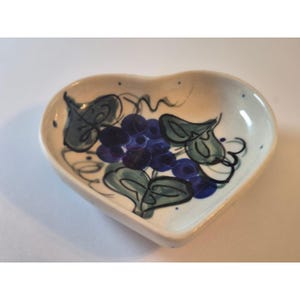 May include: Heart-shaped ceramic dish with a hand-painted design of blue grapes and green leaves. The dish has a cream-colored base and is decorated with dark blue and green accents. The dish is a decorative item.