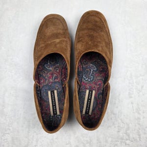 May include: A pair of brown suede loafers featuring a rounded toe and a slip-on design. The interior lining displays a colorful paisley pattern in shades of blue, red, and cream, with a white label reading Hush Puppies stitched inside each shoe.