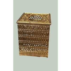 May include: A rectangular gold-colored metal tissue box cover featuring an intricate openwork lattice pattern on all sides and the top. The design includes horizontal bands of repeating diamond shapes and floral motifs with a central oval opening.