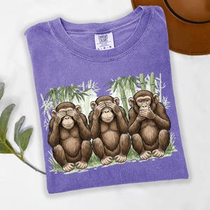 May include: A purple Comfort Colors t-shirt featuring the three wise monkeys: see no evil, hear no evil, speak no evil. The graphic shows the monkeys in front of bamboo stalks. The shirt has a crew neck and short sleeves.