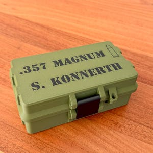 Könnte beinhalten: Ein olivgrüner, rechteckiger Aufbewahrungsbehälter mit einem schwarzen Verschluss. Auf dem Deckel befindet sich der schwarze Aufdruck .357 MAGNUM und S. KONNERTH sowie ein kleines grafisches Symbol einer Patrone. Der Behälter hat eine strukturierte Oberfläche.