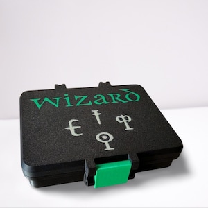 Könnte beinhalten: Schwarze rechteckige Box mit dem Wort "Wizard" in grüner Schrift. Die Box hat einen grünen Verschluss und weiße Symbole. Die Box ist geschlossen und scheint aus Kunststoff zu sein.