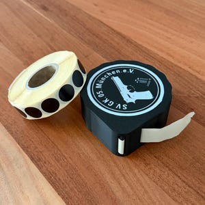 Könnte beinhalten: Eine Rolle mit kleinen schwarzen runden Aufklebern und ein schwarzer Kunststoff-Abroller. Der Abroller zeigt ein weißes kreisförmiges Logo mit einer Pistolen-Grafik und dem Text SV. GK. 05. München.e.V. auf der Oberseite.