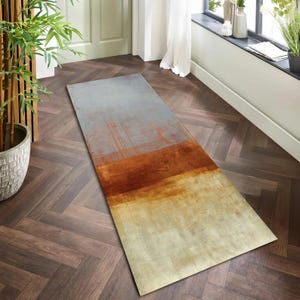 Könnte beinhalten: Ein rechteckiger Teppich mit abstraktem Design. Der Teppich weist einen Farbverlauf auf, darunter Grau, Rost und Beige. Das Design hat ein strukturiertes Aussehen mit pinselstrichartigen Mustern. Der Teppich liegt auf einem Holzboden.