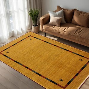 May include: Mustard yellow rectangular area rug with a border of red, black, and blue stripes. The rug is placed on a hardwood floor in a living room setting. The rug has a simple geometric design.