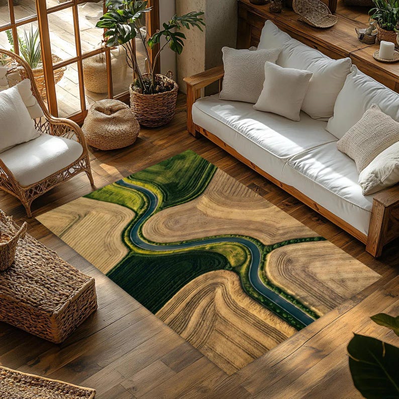 May include: Rectangular area rug with an aerial view design of a river winding through fields. The rug features shades of green, brown, and blue, creating a natural landscape pattern. The rug is placed on a wooden floor.