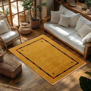 May include: A rectangular mustard-yellow rug with a dark border and decorative corner accents. The rug is placed on a hardwood floor in a living room setting, with a white sofa and wicker chair visible.