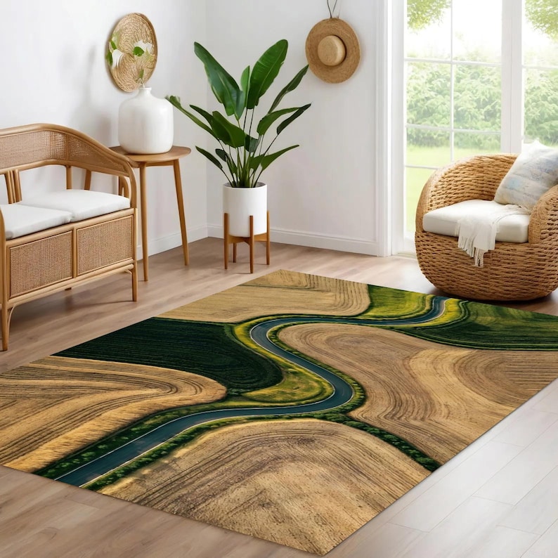May include: Area rug with a landscape design featuring a winding road through fields of brown and green. The rug has a rectangular shape and is placed on a wooden floor. The rug's design resembles an aerial view of a natural landscape.