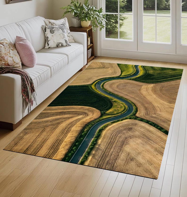 May include: Area rug featuring an aerial view of a winding road through a landscape of fields. The rug's design includes shades of brown, green, and blue, creating a naturalistic and scenic pattern. The rug is rectangular and placed on a hardwood floor.
