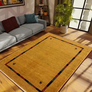 May include: A rectangular, golden-yellow area rug with a patterned border. The border features a red, blue, and black stripe design, with small black animal silhouettes in each corner. The rug is placed on a hardwood floor.