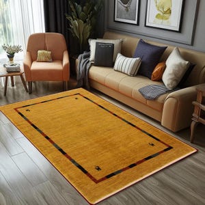 May include: A rectangular mustard-yellow rug with a dark border featuring red, black, and green stripes. The rug is placed on a hardwood floor in a living room setting, with a sofa and armchair visible in the background.