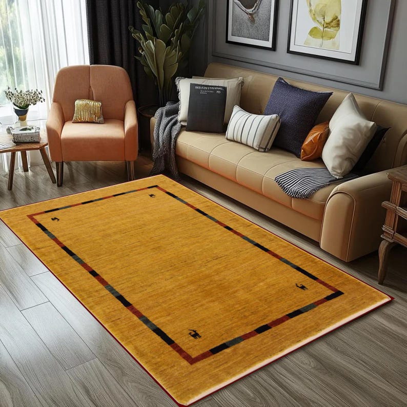May include: A rectangular mustard-yellow rug with a dark border featuring red, black, and green stripes. The rug is placed on a hardwood floor in a living room setting, with a sofa and armchair visible in the background.