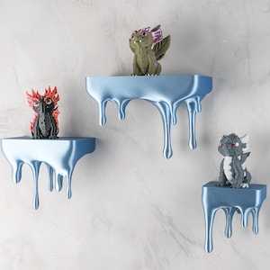 May include: Three light blue, wall-mounted shelves with a dripping paint design, each holding a small dragon figurine. The dragons are in various colors, including black with red accents, green with purple wings, and gray with white horns.
